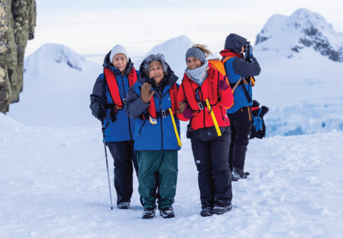Three women in winter gear and life vests pose in a snowy landscape with mountains.